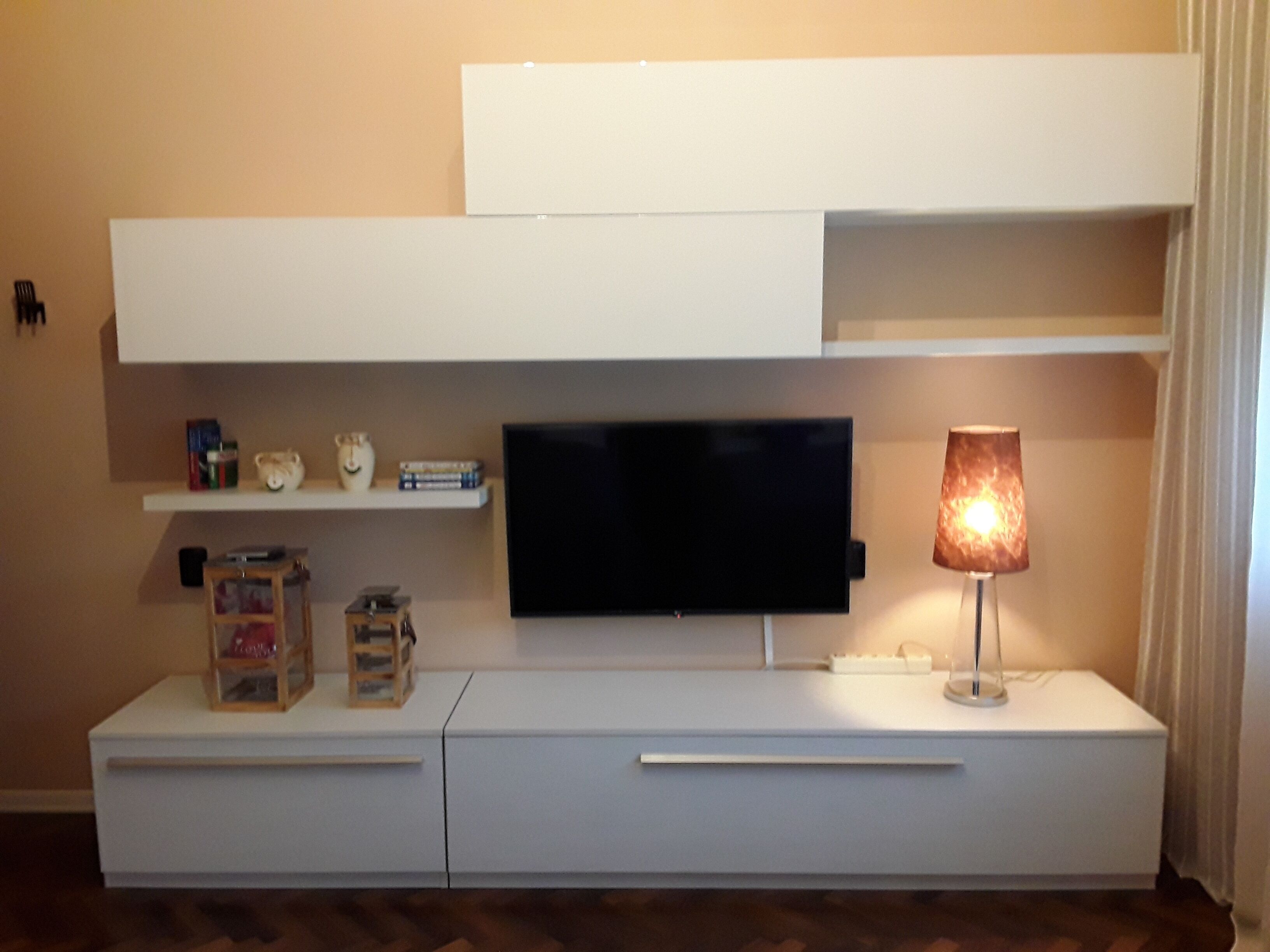 Ein modernes Wohnzimmer mit einem flachbildfernseher an einer weißen Unterhaltungseinheit, einem kleinen Tisch mit Lampe und einem Regal mit Büchern und Dekorationsgegenständen.