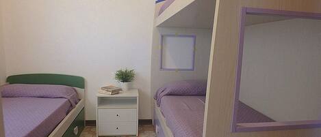 Une chambre avec deux lits superposés, une petite table de chevet ornée d’une plante et de quelques livres, et un tableau encadré accroché au mur.