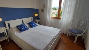 Un dormitorio con cama, dos mesitas de noche, una silla, un ventanal con cortinas y vistas al exterior.