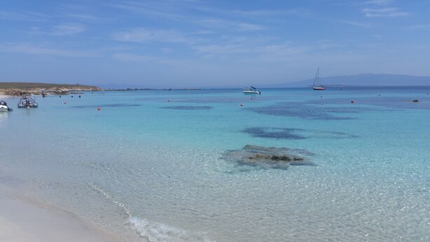 Una playa con agua turquesa cristalina, barcos y afloramientos rocosos.