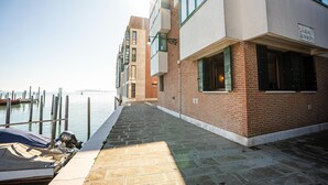 Een gebouw aan het water met een bakstenen gevel, grote ramen en een verharde looppad naar een steiger met boten.