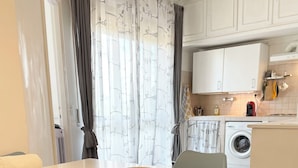 Zona pranzo con tavolo bianco e sedie blu, lavatrice e cucina con mobili bianchi e forno a microonde.