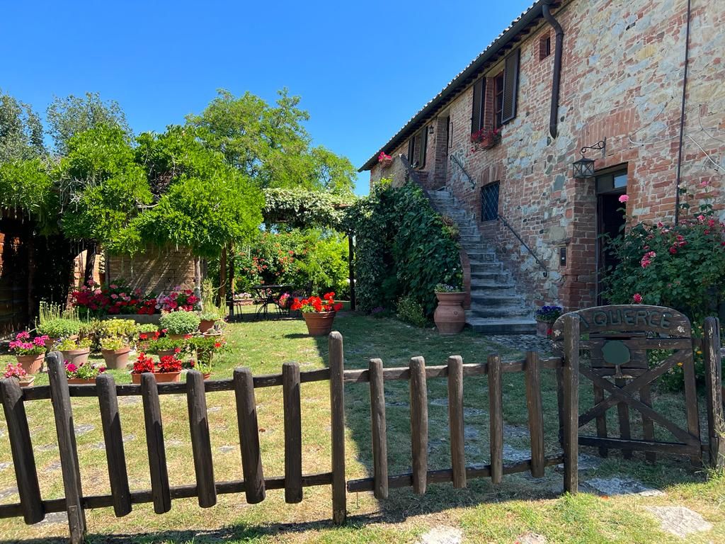 A rustic stone building with a wooden fence and a gate, surrounded by lush greenery and potted plants.