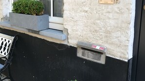 En byggnad med en vit fasad, en svart nedre del, ett fönster med flera rutor, en liten blomkruka med gröna växter, en metallporttelefon och en bänk.