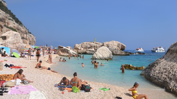 Una playa con gente tomando el sol y nadando, formaciones rocosas y barcos en el agua.