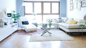 A modern living room with a white sofa, a glass coffee table, and a leather pouf. There is a large window with wooden shutters, a white lamp, and a plant on a side table. - Beautiful, Cosy, Great Apartment Downtown, Near Beach.  Ask for Parking. (Alicante)