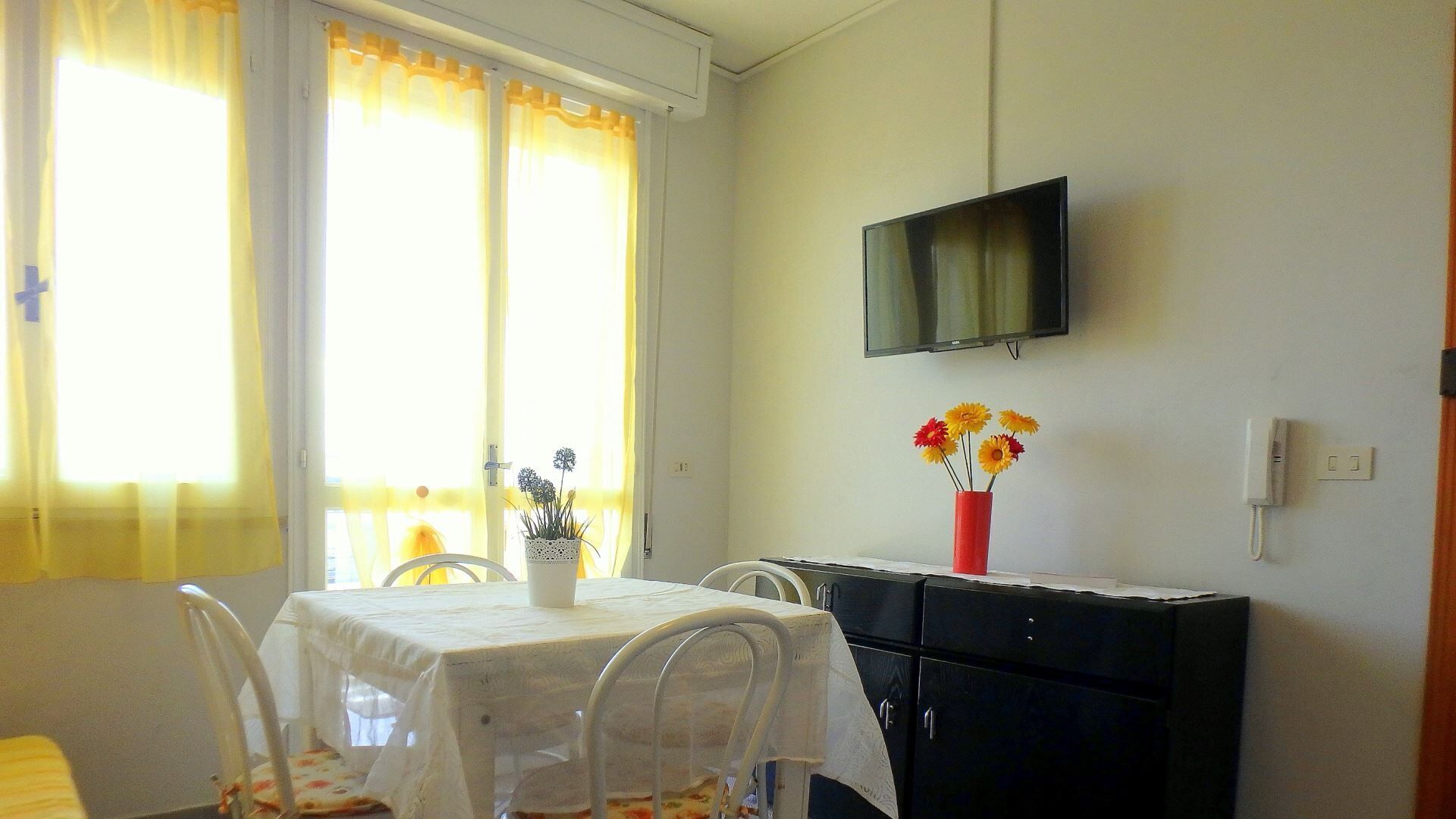 A dining area with a table set for two, a TV mounted on the wall, and a vase with flowers on a cabinet.