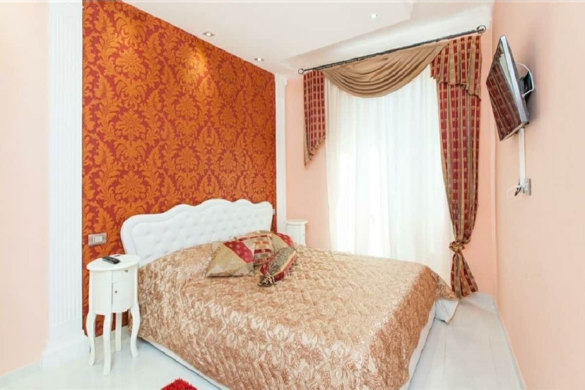 A bedroom with a patterned wallpaper, a white headboard, a bed with a patterned bedspread, a bedside table, and a TV mounted on the wall.