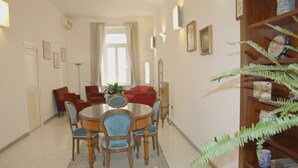 A dining area with a round table, chairs, and a patterned rug. There is a bookshelf with decorative items and a potted plant. - Luxurious apartment in an ancient noble palace in the historic center of Naples (Napoli)