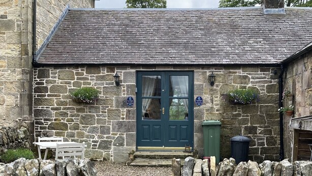 A stone cottage with a dark blue door, a small white table, and a stone wall in front. - Charming Rural Farmhouse Cottage (by Carnwath)