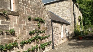 A stone building with a brick wall and a small garden area with potted plants. - Charming Rural Farmhouse Cottage (by Carnwath)