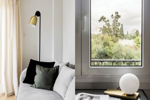 Ett modernt vardagsrum med en soffa, en golvlampa och utsikt över en balkong med grönska och byggnader.