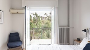 A bedroom with a bed, a chair, a radiator, and a view of trees and buildings through an open door. - UPSTREET Zeus Temple View 2BD Apt Acropolis-Plaka (Athens)