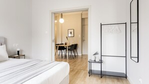 A hotel room with a bed, a small table with a lamp, a chair, a dining area with a table and chairs, and a coat rack. - UPSTREET Zeus Temple View 2BD Apt Acropolis-Plaka (Athens)