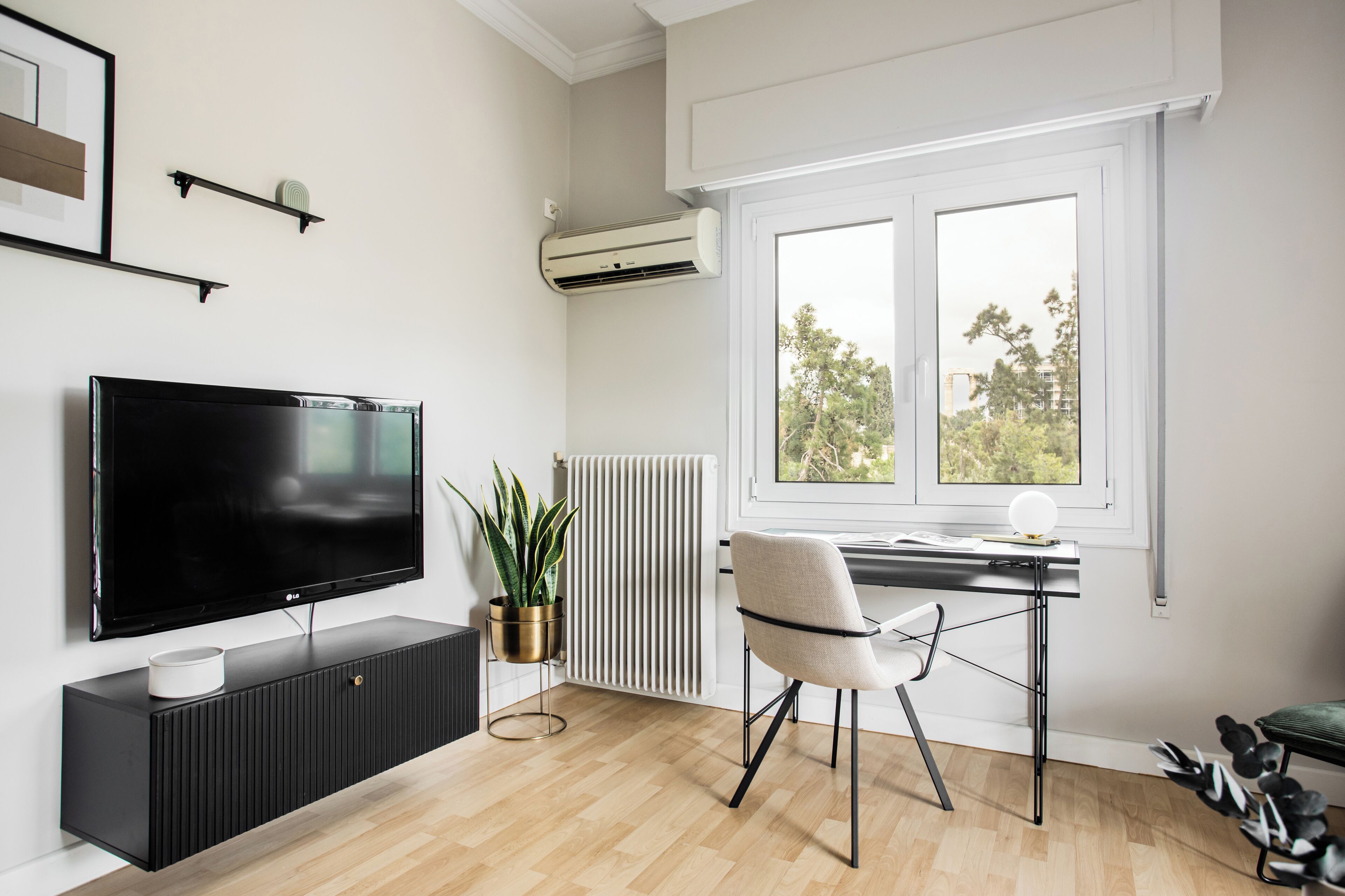 A modern living room with a flat-screen TV, a radiator, a potted plant, a chair, and a window with a view of trees.