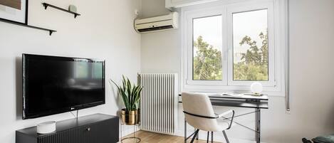A modern living room with a flat-screen TV, a radiator, a potted plant, a chair, and a window with a view of trees.