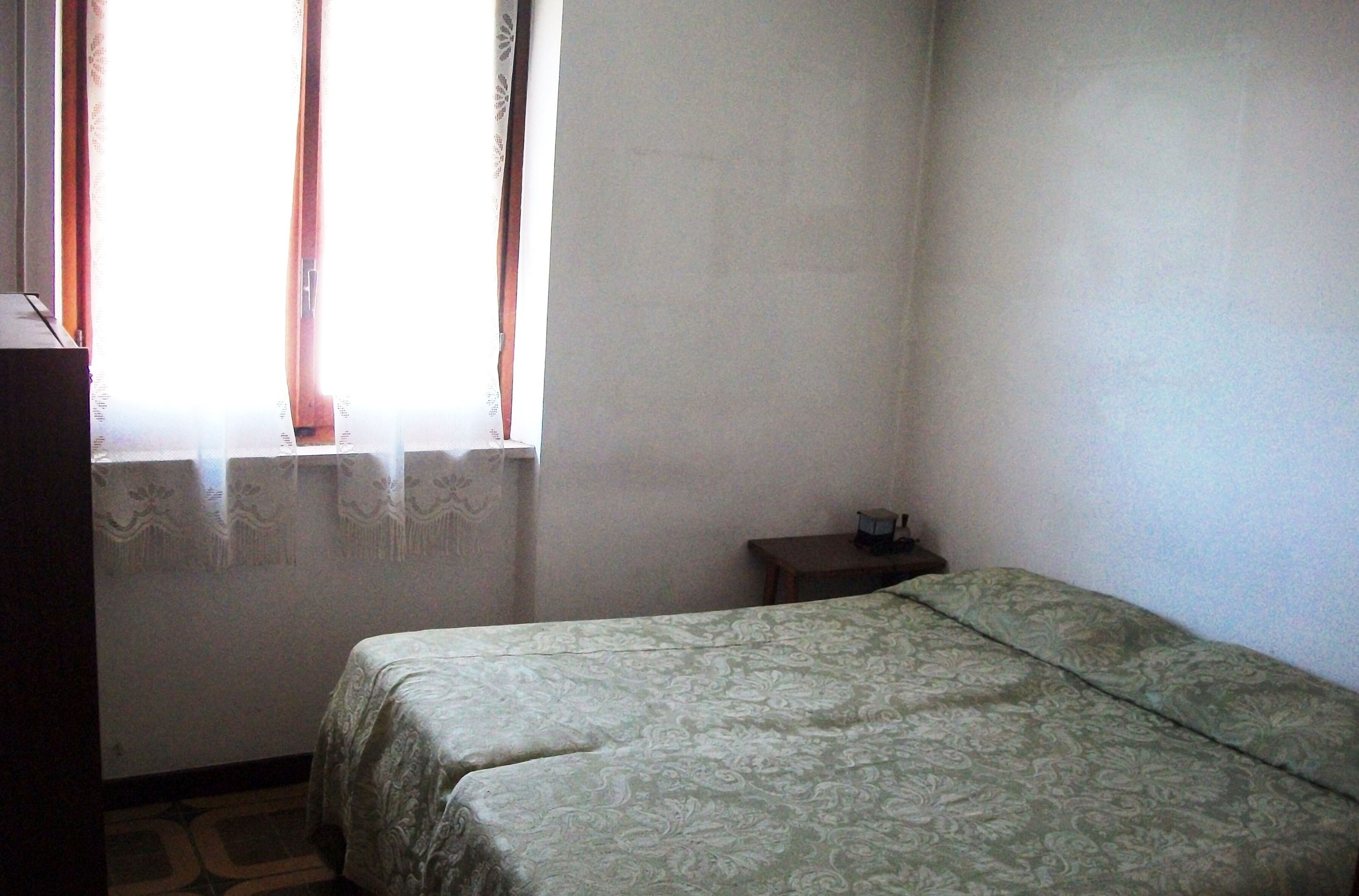 Ein kleiner, schlichter Raum mit einem Bett, einem kleinen Tisch und einem Fenster mit Vorhängen.