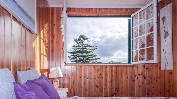 A bedroom with wooden walls, a bed with purple and white bedding, a window with a view of a tree, and a painting on the wall.