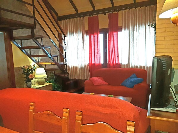 Une pièce avec un canapé rouge, une chaise en bois, un téléviseur posé sur un meuble, une lampe et un escalier.