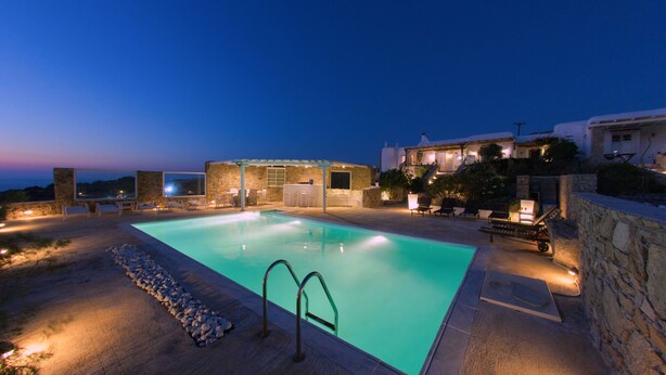 Una piscina con borde de piedra y escalera de metal, rodeada de sillones de descanso y un muro de piedra.
