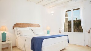 Un dormitorio con cabecera de madera, ropa de cama blanca, un plaid azul, dos lámparas de noche y vistas a través de una ventana.