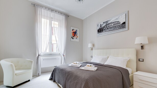 Quarto de hotel moderno com uma cama grande, uma poltrona branca, uma janela com cortinas leves e obras de arte nas paredes.