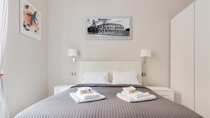 Quarto de hotel com uma cama grande, duas mesinhas de cabeceira brancas, cabeceira e um quadro com imagem do Coliseu.