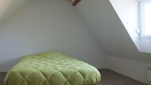 A room with a sloped ceiling, a wooden beam structure, a green upholstered bed, and a window providing natural light.