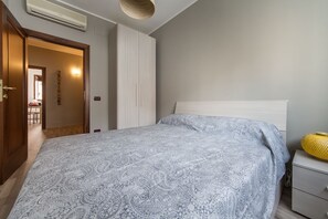 A bedroom with a bed, a bedside table, a wall-mounted air conditioner, and a door leading to another room.