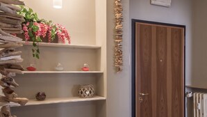 A hallway with wooden flooring, a wooden door, and a radiator. Shelves with decorative items and a hanging light fixture are visible.