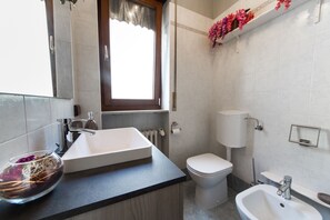 A modern bathroom with a large white sink, a toilet, and a bidet. There is a window with a curtain, and a shelf with flowers above the toilet.