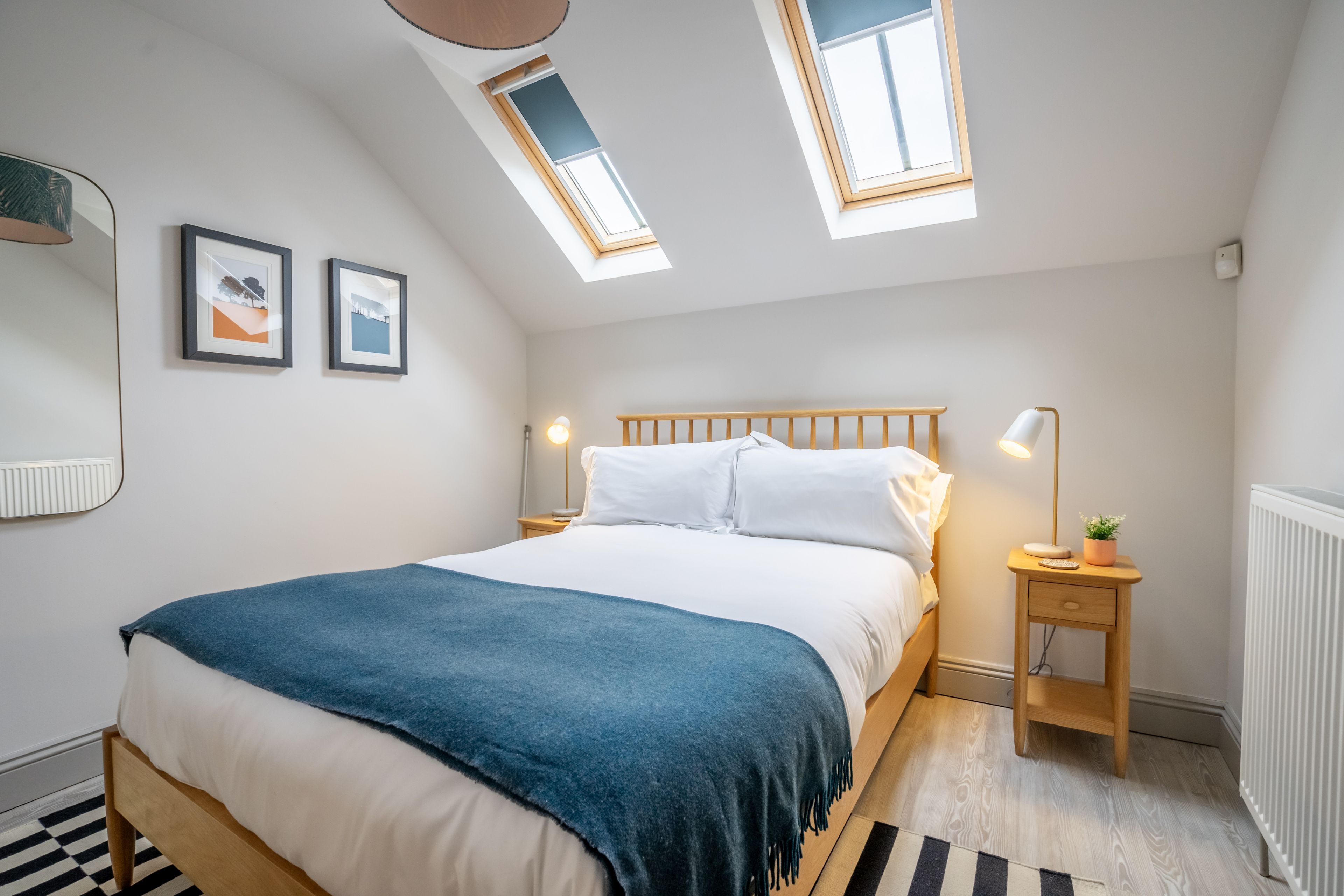 A bedroom with a bed, skylights, a bedside table with a lamp, and framed pictures on the wall.