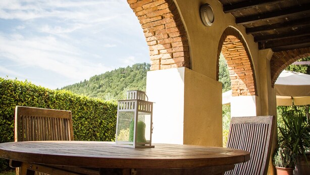Un patio exterior con una mesa redonda de madera, dos sillas de madera, un farol y un arco de ladrillo.