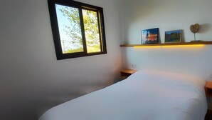 Une chambre à coucher avec un lit, une fenêtre donnant sur des arbres, une étagère avec deux tableaux encadrés et une table de chevet.