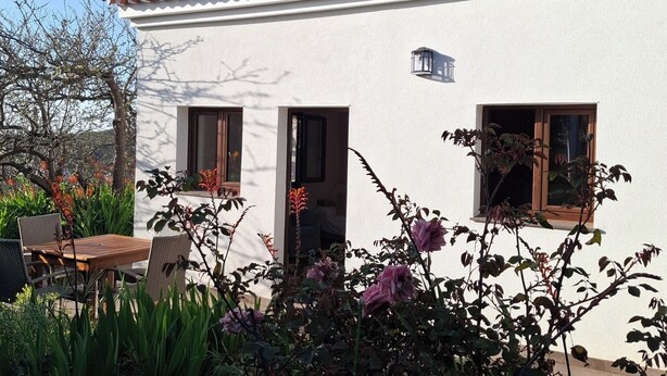 Un bâtiment blanc avec un toit recouvert de tuiles, deux fenêtres et une porte. On y trouve une table et des chaises à l’extérieur, entourées de plantes et de fleurs.