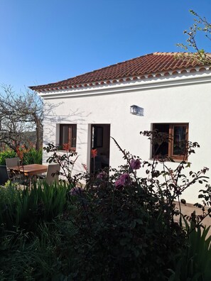 A white building with a tiled roof, two windows, and a door. There is a table and chairs outside, surrounded by plants and flowers.