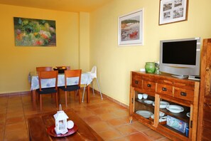 Un coin repas avec une table dressée pour deux personnes, des chaises, un téléviseur posé sur un meuble en bois et un sol carrelé.