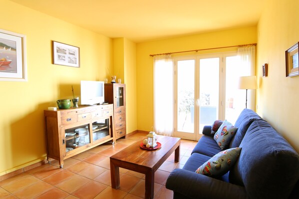 Un salon avec un canapé, une table basse en bois, un buffet contenant de la vaisselle et une fenêtre avec des rideaux.