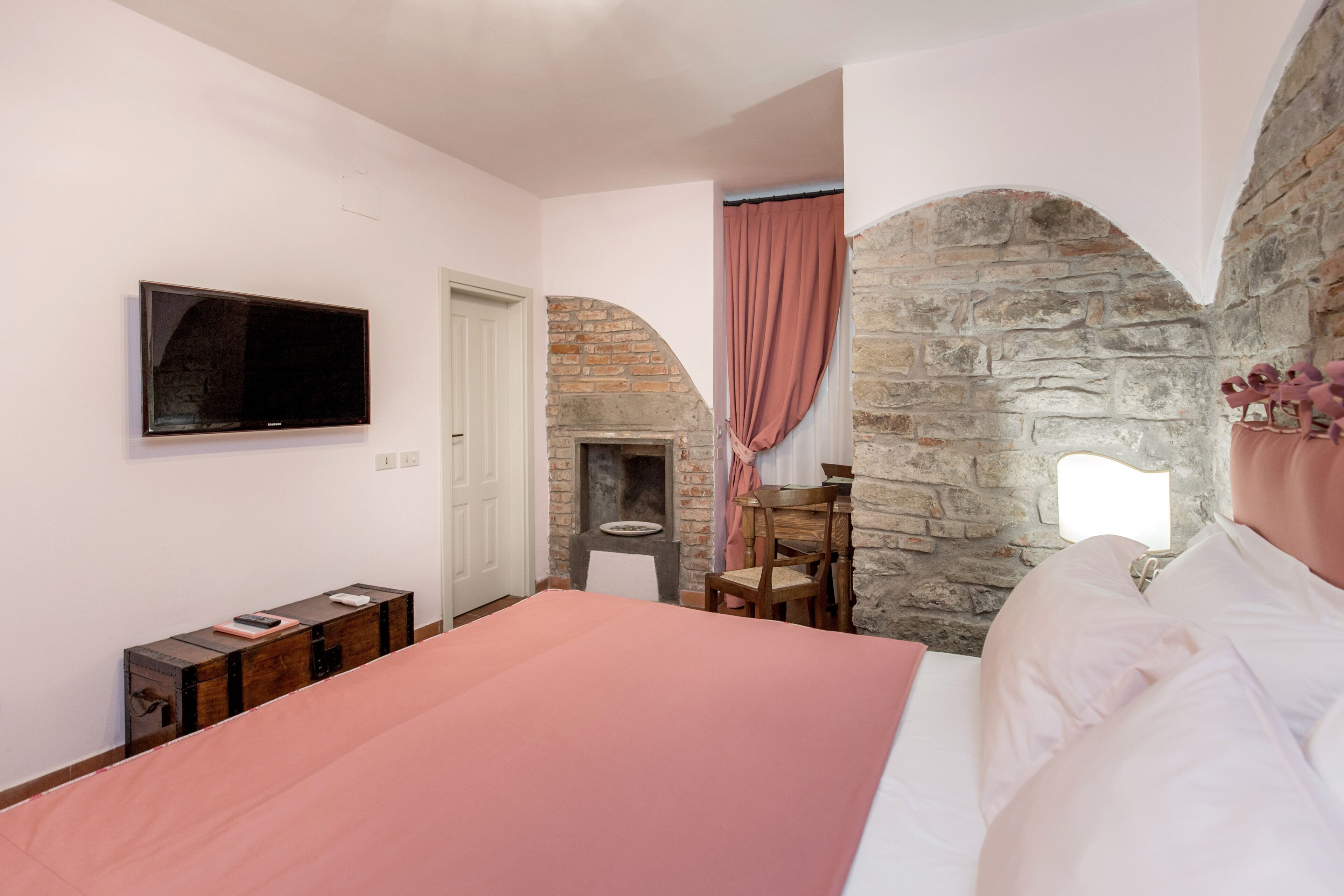 Une chambre avec une cheminée en pierre, un téléviseur fixé au mur, un lit recouvert d’une couverture rose et une chaise en bois.