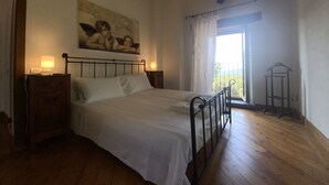 A bedroom with a bed, wooden floor, a nightstand, a wall-mounted lamp, a painting, and a window with curtains. -  Authentic Massa Marittima Get Away 220 sqm in the Tuscan countryside  (Massa Marittima)