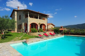 A stone villa with a swimming pool, red shutters, and a view of the mountains.