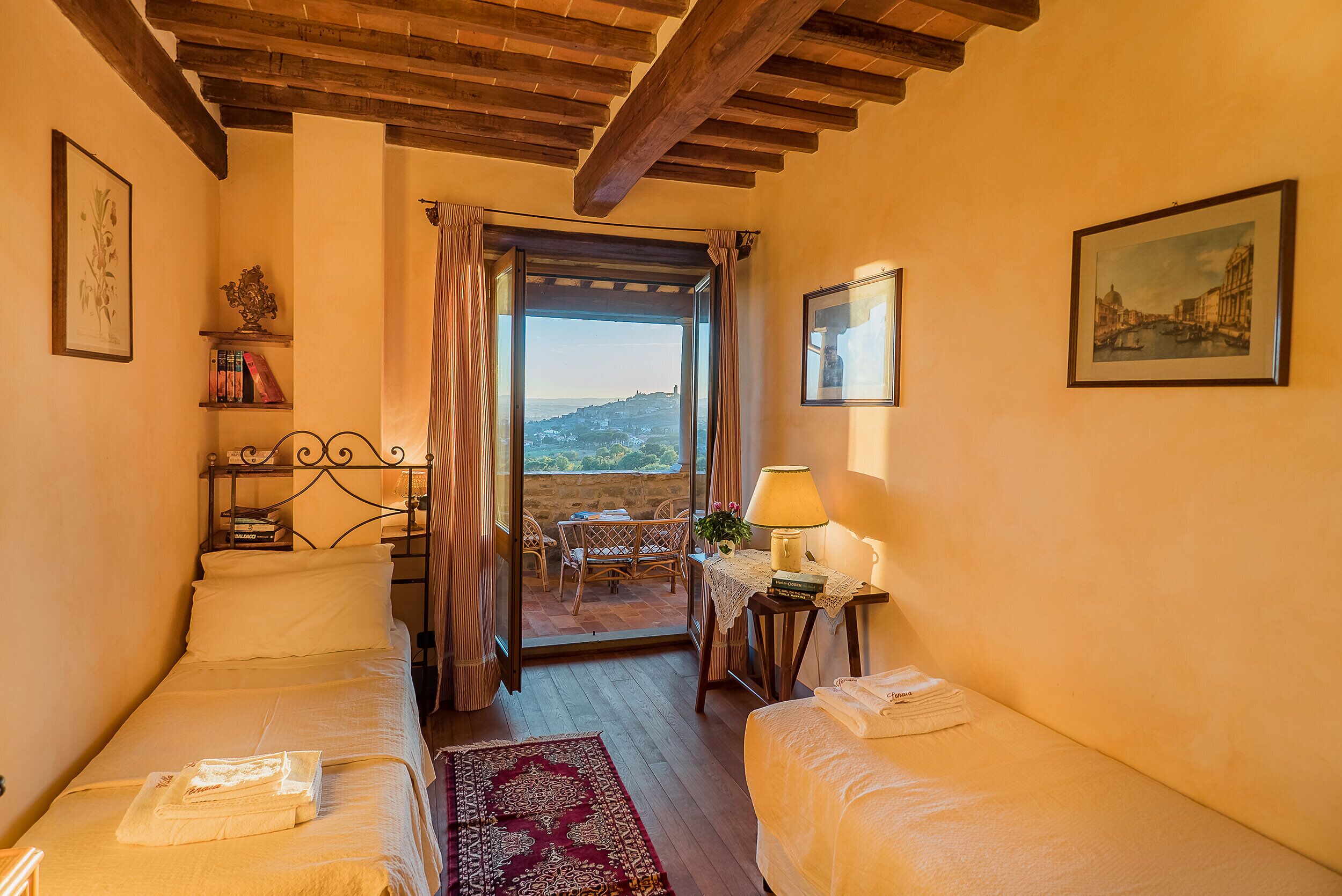 A cozy room with two beds, a table with a lamp, a view of a balcony, and a painting on the wall.