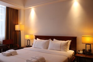 Ein Hotelzimmer mit einem großen Bett, zwei Nachttischlampen, einem Schreibtisch mit Stuhl und einem Fenster mit Vorhängen.