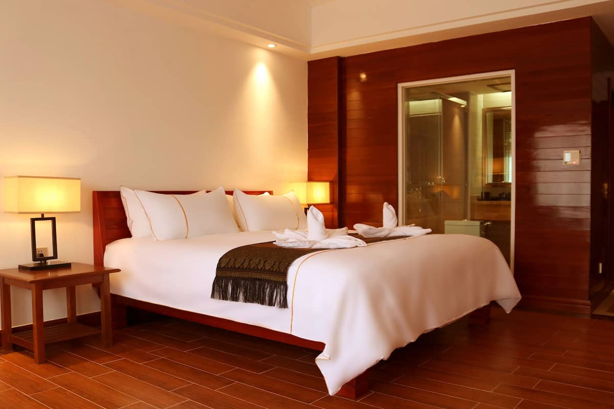 Een hotelkamer met een groot bed, twee bedlampjes, een houten hoofdeinde, een houten vloer en een glazen deur naar een badkamer.