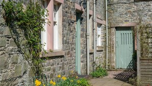 A stone building with a narrow stone pathway, a green door, and yellow flowers along the path.