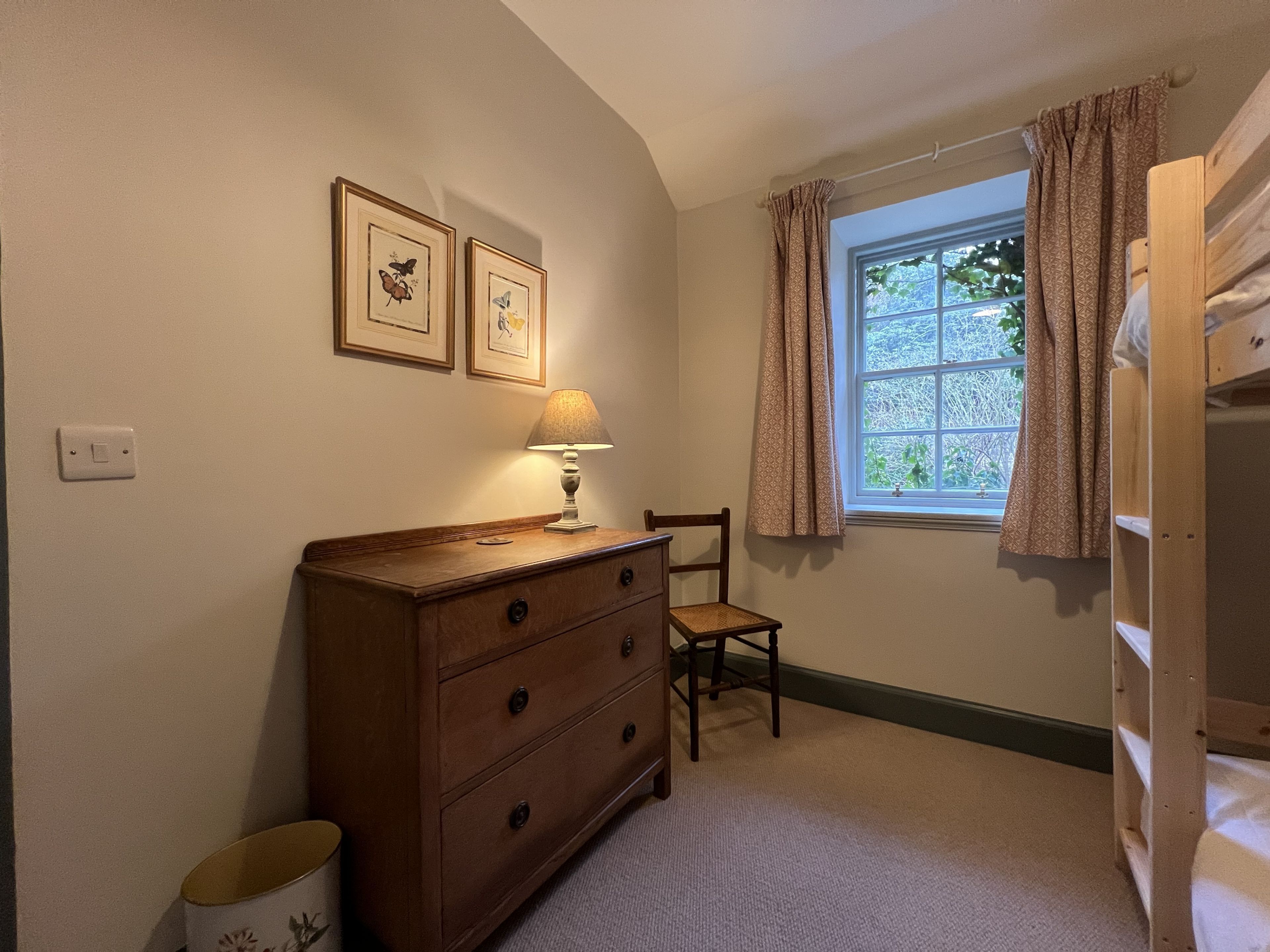 Ein Schlafzimmer mit einem Holzkoffer, einem Sessel, einem Fenster mit gemusterten Vorhängen und gerahmten Schmetterlingsbildern an der Wand.