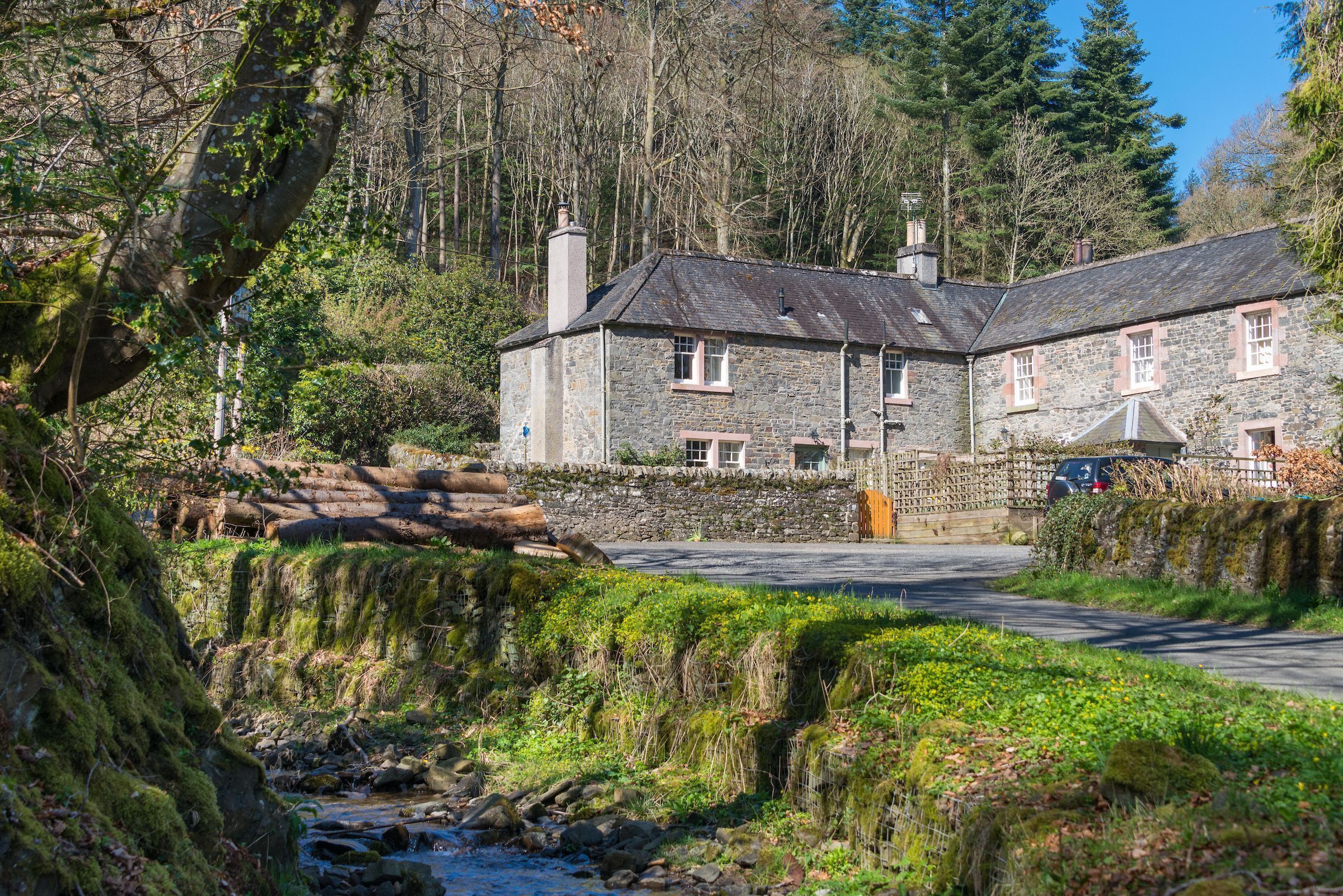 A stone building with a stone roof, surrounded by greenery and a stream.