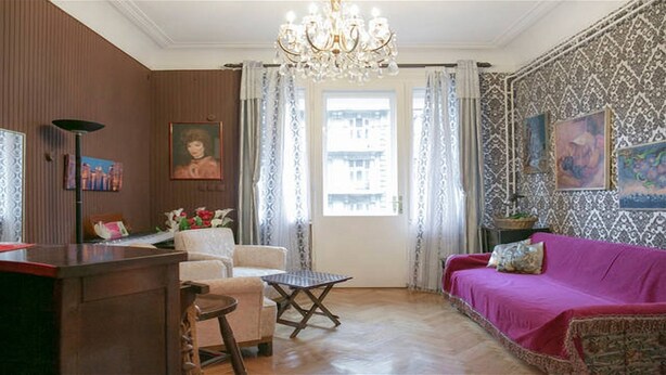 Ein Zimmer mit Holzboden, einem rosafarbenen Sofa, einem Holzstuhl, einem kleinen Tisch, einer Chandelier und Wandtapete mit Muster.