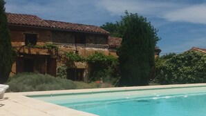 Vista de una piscina con un edificio de techo a ladrillo, rodeado de follaje y un cielo despejado.