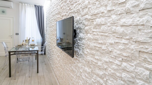 Un comedor con una mesa y sillas de vidrio, una pared de piedra y un televisor montado.
